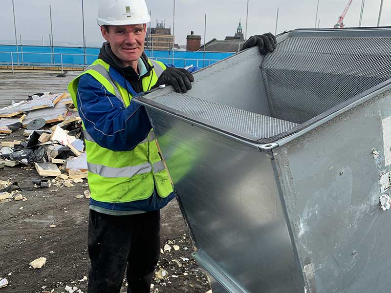 close up photo of a howarth recycling scrap collections expert dressed in blue sleeves poking out from a hi vis yellow vest posing next to a very large aluminimium air shaft square
