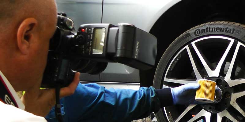 clos up behind angled view of paul roberts from itworknplay photographing a vehicle fuel filter in front of a car wheel held by one of excel automotives garage mechanics